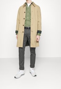 Beige trenchcoat med bruna knappar över en grön skjorta, grå byxor och vita sneakers. Rutigt foder synligt vid ärmarna.