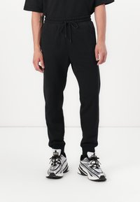 Black, tapered jogger pants with an elastic waistband and drawstring. Paired with gray and black athletic shoes featuring white accents.