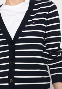 Marinblå kofta med vita horisontella ränder, V-ringning, knäppning med guldfärgade knappar och kontrasterande logodetalj på bröstet.