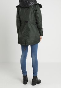 Giacca parka verde con cappuccio trapuntato nero, coulisse in vita e cerniere laterali, abbinata a jeans blu slim e stivaletti neri alla caviglia.