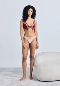 Μπορντό σουτιέν με δαντέλα και κυματιστά χείλη, συνδυασμένο με ροζ εσώρουχο από δαντέλα. Το μοντέλο στέκεται σε απαλό γκρι χαλί κοντά σε λευκό καμπύλο καναπέ.