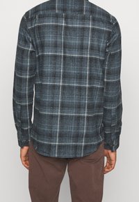 Camicia a quadri blu con maniche lunghe, realizzata in tessuto testurizzato. Linee bianche creano un motivo a scacchi. Presenta polsini con bottone e orlo curvo.