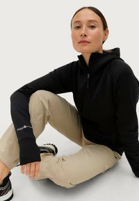 Zwarte zip-up jas met een hoge kraag, gedragen over beige broeken. De jas heeft lange mouwen en een logo op de manchetten.