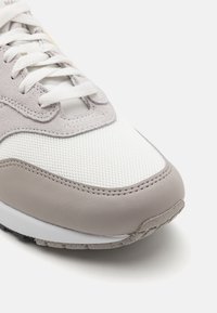 Grå og hvid sneaker med mesh- og ruskindsmaterialer, rund tå, flade snørebånd og struktureret gummisål med sprøjtede accenter.