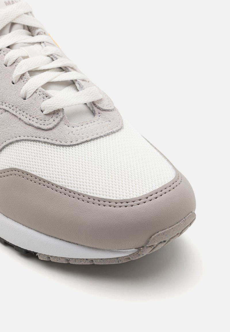 Zapatilla gris y blanca que presenta materiales de malla y ante, puntera redonda, cordones planos y suela de goma texturizada con acentos moteados.