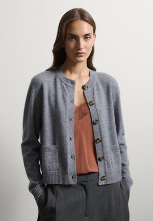 Mujer con cárdigan gris abotonado sobre camisola de encaje en color óxido y pantalones gris oscuro, de pie con las manos en los bolsillos contra un fondo liso.