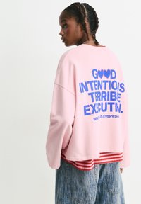 Felpa rosa con scritta blu sulla schiena che recita "BUONE INTENZIONI, ESECUZIONE TERRIBILE." Indossata sopra una maglietta a righe rosse, abbinata a una gonna di jeans.
