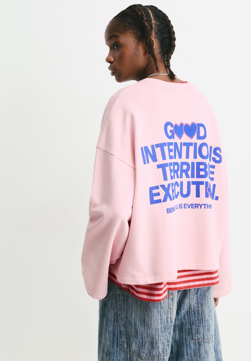 Felpa rosa con scritta blu sulla schiena che recita "BUONE INTENZIONI, ESECUZIONE TERRIBILE." Indossata sopra una maglietta a righe rosse, abbinata a una gonna di jeans.