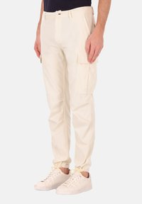 Crème-kleurige cargobroeken met zijzakken, taps toelopende pijpen en elastische boorden, gemaakt van zachte stof. Gedragen met lichte sneakers.