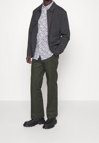 Tummanharmaa takki, kukkakuvioinen valkoinen ja sininen paita, oliivivihreät housut, mustat paksupohjaiset saappaat. Sileä kangas, rakenteellinen istuvuus, napitus takissa.