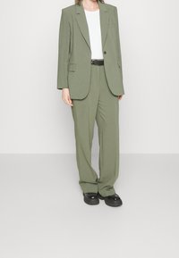 Completo sartoriale verde oliva con blazer strutturato e pantaloni a gamba larga, abbinato a una t-shirt bianca e una cintura nera, completato da scarpe nere.