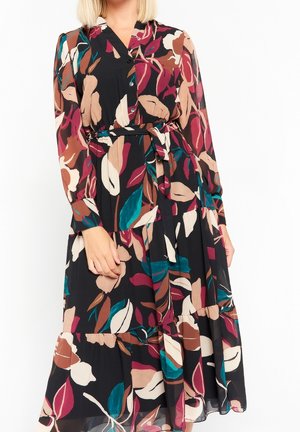 Donna che indossa un vestito a maniche lunghe con bottoni e cintura, caratterizzato da un motivo astratto di foglie multicolori in nero, turchese, rosa, marrone e beige.