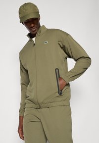 Λαδί πράσινη ζακέτα με φερμουάρ και ψηλό γιακά, ελαστικούς καρπούς και τσέπες με φερμουάρ. Συνδυάζεται με ταιριαστά παντελόνια και καπέλο, με λογότυπο.