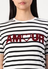 Svart-hvite stripete t-skjorte med røde, teksturerte "AMOUR" bokstaver og hjerte-motiv, rund hals, korte ermer.