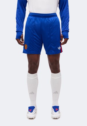 Männlicher Sportler, der blaue Manchester United-Shorts, weiße knielange Socken, weiße Fußballschuhe und ein blau-weißes Langarmoberteil trägt, steht aufrecht.