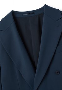 Blazer doppiopetto blu navy con revers a punta, texture liscia e rivestimento interno visibile sul retro. Dettaglio di una tasca sul petto evidente.