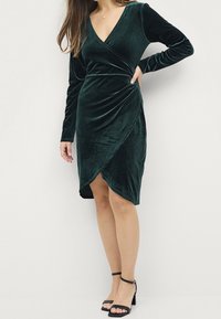 Dunkelgrünes Samtkleid mit langen Ärmeln, V-Ausschnitt und Wickeldesign, das ein gerafftes Seitendetail aufweist. Kombiniert mit schwarzen Riemchen-High Heels.