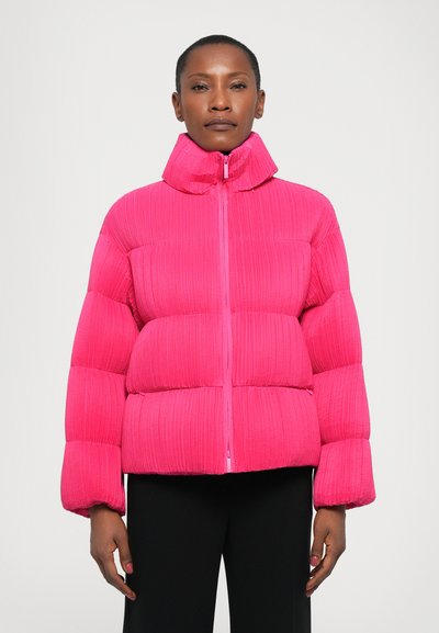 Frau in einer leuchtend rosa, gesteppten Daunenjacke mit hohem Kragen, steht vor einem schlichten weißen Hintergrund, die Hände entspannt an den Seiten.