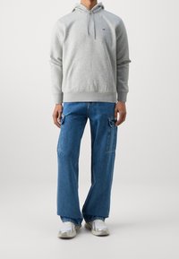 Felpa grigia con tasca frontale e cappuccio con cordino, abbinata a jeans blu a vestibilità ampia con tasche laterali. Sneakers bianche.