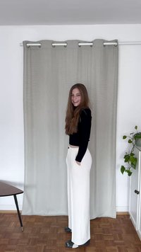 White wide-leg trousers with pockets, a black fitted long-sleeve top, and grey shoes. Background features a light grey curtain and plant.