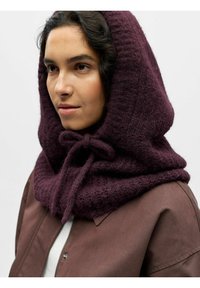 Écharpe à capuche en tricot violet avec un motif texturé et un détail à nouer, portée sur une veste marron. Le tissu semble doux et chaud.