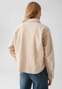 Woman with long blond hair wears a loose beige corduroy jacket and blue jeans, shown from behind against a plain background.