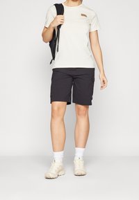 Světle béžové tričko s malým hnědým logem, v kombinaci s černými cargo šortkami. Osoba má na sobě smetanové tenisky a bílé ponožky, nese černý batoh.