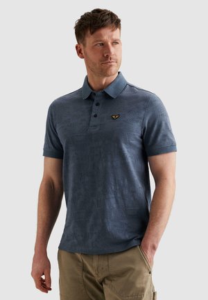 Man wearing blue patterned polo shirt with small embroidered logo and khaki pants, standing with one hand in pocket.