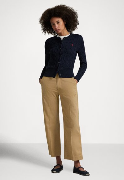 Γυναίκα με σγουρά μαλλιά που φοράει ναυτικό μπλε πουλόβερ με πλεκτό σχέδιο και μπεζ παντελόνι φαρδύ στο πόδι, καθώς και μαύρα παπούτσια Mary Jane, στέκεται με τα χέρια στις τσέπες.