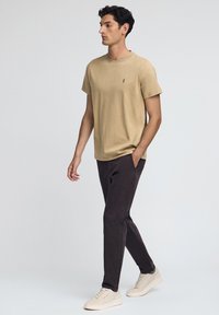 Beżowa bawełniana koszulka z okrągłym dekoltem i małym logo, w połączeniu z ciemnobrązowymi spodniami i beżowymi trampkami. Model stoi w profilu.