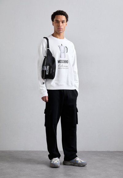 Sudadera blanca de manga larga con estampado gráfico, pantalones cargo negros y zapatillas grises con acentos coloridos. Mochila cruzada negra con logo.