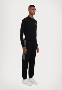 Polo nero con maniche lunghe e colletto, abbinato a pantaloni jogger neri con dettagli grigi sui lati. Sneakers bianche completano l'outfit.