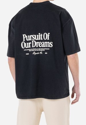 Osoba nosząca czarną koszulkę z białym napisem „Pursuit Of Our Dreams” oraz dodatkowym małym tekstem na plecach, w połączeniu z jasnymi spodniami.