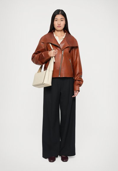 Giacca da biker in pelle marrone con ampio colletto, chiusura con zip e maniche larghe. Abbinata a pantaloni neri a gamba larga e una borsa a spalla beige.