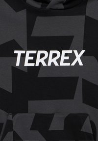 Μαύρο και σκούρο γκρι ύφασμα με μοτίβο και λευκό κείμενο "TERREX" τυπωμένο στο μπροστινό μέρος πάνω από μια τσέπη καγκουρό.