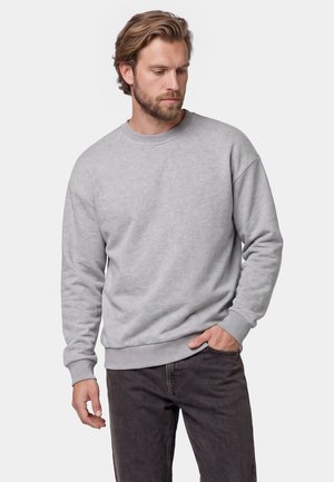 Mand med lyst brunt hår og skæg iført en lysegrå sweatshirt og mørke jeans, stående med den ene hånd i lommen, kiggende ned til siden.