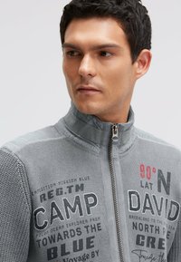 Graue Jacke mit Reißverschluss, die strukturierte Strickärmel, einen glatten Körper und auffällige bedruckte Grafiken in Schwarz und Rot aufweist.