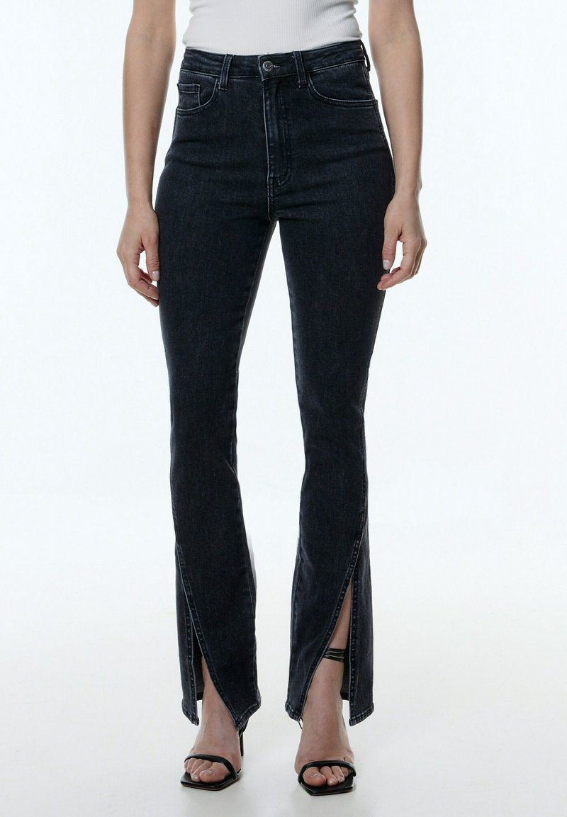 Femme portant un jean noir taille haute avec des fentes à l'avant des chevilles, associé à des sandales à talons noires à brides et un top blanc sans manches.