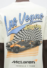 Krēmkrāsas T-krekls ar sacīkšu automašīnas grafiku, šaha karoga rakstu un tekstu “Las Vegas Racing” zilā un dzeltenā krāsā. Iekļauts McLaren logotips.