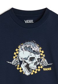 Camiseta de algodón azul marino con un diseño gráfico que presenta una calavera, olas y un patrón de tablero de ajedrez en amarillo y azul. Logotipo de "VANS" exhibido.