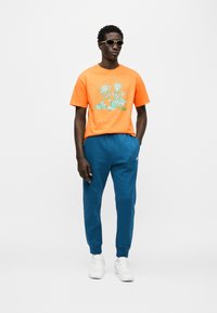 Πορτοκαλί μπλουζάκι με γραφικό σχέδιο, μαύρο παντελόνι και λευκά αθλητικά παπούτσια. Αξεσουάρ περιλαμβάνουν γυαλιά ηλίου και λογότυπο στα ρούχα. Καθημερινή εμφάνιση.