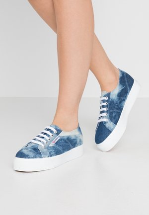 Jambes portant des baskets compensées en denim tie-dye bleu avec des lacets blancs et des semelles épaisses blanches sur un fond clair uni.