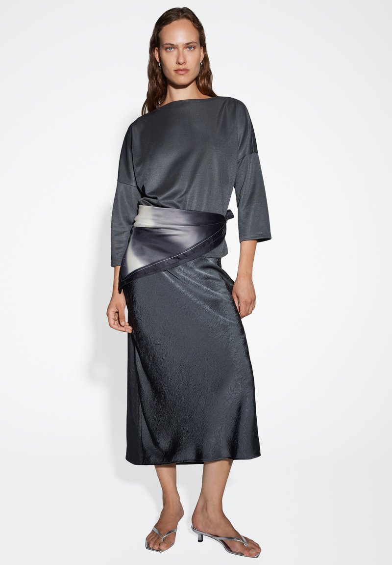 Top grigio scuro, simile a raso, con una vestibilità ampia e maniche a tre quarti, abbinato a una gonna lunga texturizzata con una fascia in gradiente scuro.