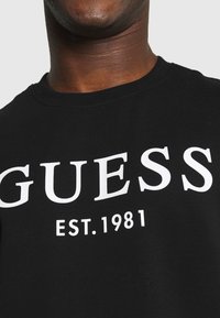 Camiseta de algodón negra con gran texto blanco "GUESS" y "EST. 1981" en la parte frontal. Cuello redondo acanalado con textura suave.