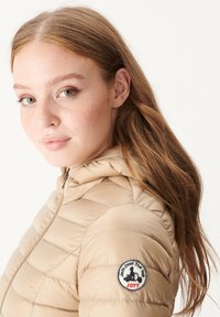 Beige gewatteerde Jas met capuchon, met een gewatteerd ontwerp, een ritssluiting aan de voorkant en een rond geborduurd logo op de linker mouw.
