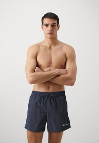 Șorturi de baie bleu marin cu talie elastică și șnur. Prezintă un mic logo alb pe piciorul stâng. Material ușor, cu textură.