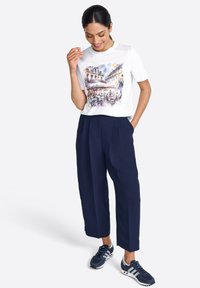 T-shirt blanc avec une scène illustrée colorée, associé à un pantalon bleu marine et des chaussures de sport bleues marines. Coupe décontractée et texture de tissu lisse.