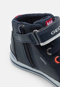 Zapato de caña alta azul marino con un exterior de cuero suave, ojales naranjas en contraste, cierre de cremallera y un collar acolchado para mayor comodidad.