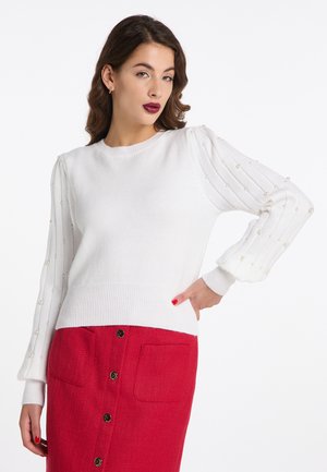 Donna con capelli castani ondulati indossa un maglione bianco a maniche lunghe con dettagli in perle e una gonna rossa con bottoni e tasche frontali.