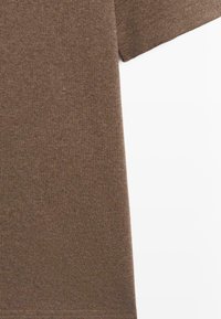 Detailní záběr na krátkosleevě hnědou látkovou košili zobrazující texturu a šití na rukávu a spodním okraji na bílém pozadí.